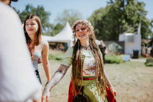 Zwei Besucherinnen des Elbenwald Festivals genießen die Atmosphäre. Eine Frau mit Locken und Blumenkranz trägt ein traditionelles Outfit, während die andere in einem lässigen Top strahlt. Im Hintergrund sind Festivalzelte und eine grüne Wiese zu sehen.