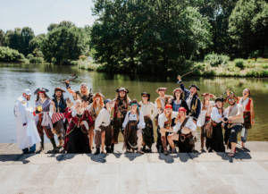 Gruppe von Cosplayern in Piratenkostümen am Ufer eines Gewässers, lächelnd und posierend für ein Foto, während sie Requisiten wie Schwerter und Kanonen halten. Die Szene spiegelt die lebendige Atmosphäre des Elbenwald Festivals wider.