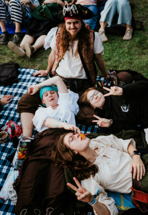 Vier Freunde in Kostümen entspannen auf einer Decke beim Elbenwald Festival. Der Pirat mit Hut lächelt, während die anderen sich mit Snacks vergnügen und peace-Zeichen zeigen. Die fröhliche Atmosphäre und die bunte Kleidung spiegeln die kreative Gemeinschaft wider.