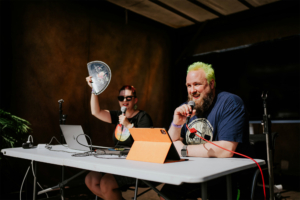 Tobias M. Eckrich Zwei Moderatoren auf der Bühne des Elbenwald Festivals, mit Mikrofonen in der Hand. Die Frau mit der bunten Frisur hält einen Fächer hoch, während der Mann mit grünem Haar lächelt. Im Hintergrund sind technische Geräte und ein entspannter Festival-Atmosphäre zu sehen.