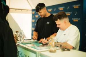 Anbu Studios Zwei männliche Fans signieren Autogramme am Elbenwald Festival. Im Vordergrund sind ein Wasserflasche und eine Glasflasche mit Dekoration sichtbar. Der Hintergrund zeigt das Festival-Logo auf einem Banner.