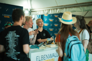 Distrikt 14 Besucher interagieren am Elbenwald Festival-Stand, umgeben von freundlichen Mitarbeitern. Die Atmosphäre ist lebhaft und einladend, während die Teilnehmer sich über das Festival und seine Angebote austauschen.