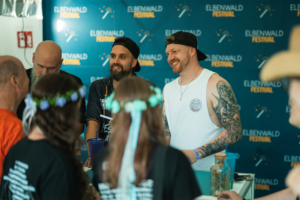 Punk Rock Factory Menscheninteraktion am Elbenwald Festival: Besucher stehen an einem Stand, während Mitarbeiter lächeln und sich mit den Fans austauschen. Im Hintergrund ist das Festival-Logo prominent zu sehen, was die freundliche und einladende Atmosphäre unterstreicht.
