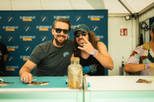 dArtagnan Zwei Männer lächeln vor einem Hintergrund mit dem Logo des Elbenwald Festivals. Sie tragen Sonnenbrillen und zeigen das "Rock on"-Zeichen. Auf dem Tisch liegen Autogrammkarten, während eine Glasvase mit bunten Gegenständen im Vordergrund steht.