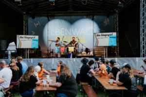 Bingo by Quizlabor Bühne beim Elbenwald Festival mit Moderatoren, die ein Bingospiel präsentieren. Im Vordergrund sitzen Festivalbesucher an Tischen und genießen Getränke, während sie gespannt dem Geschehen auf der Bühne folgen. Atmosphäre von Spaß und Gemeinschaft.
