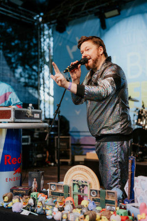 Bingo by Quizlabor Ein Moderator in einem glitzernden Anzug spricht ins Mikrofon auf der Bühne des Elbenwald Festivals, umgeben von bunten Merchandise-Artikeln und Spielzeugen. Im Hintergrund sind Musik-Equipment und eine festliche Atmosphäre zu sehen.