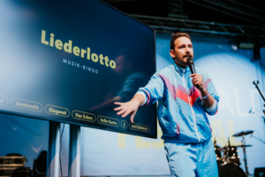 Bingo by Quizlabor Ein Moderator präsentiert das Liederlotto, ein Musik-Bingo-Spiel, auf der Bühne des Elbenwald Festivals. Im Hintergrund ist eine große Bildschirmpräsentation mit Spielanweisungen zu sehen. Das Event vereint Musik und Nerd-Kultur in einer lebhaften Atmosphäre.
