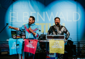 Bingo by Quizlabor Zwei Moderatoren auf der Bühne des Elbenwald Festivals, gekleidet in auffälligen Outfits, präsentieren ein Quiz-Event. Im Hintergrund ist das Festival-Logo sichtbar, während bunte Flaggen mit dem Schriftzug "Quizlabor" dekorativ auf dem Tisch hängen.