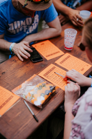 Bingo by Quizlabor Teilnehmer des Elbenwald Festivals sitzen an einem Tisch und spielen ein Gesellschaftsspiel. Auf dem Tisch liegen orangefarbene Spielblätter, ein Smartphone und eine Tüte mit bunten Spielsteinen. Die entspannte Atmosphäre spiegelt die kreative Stimmung des Festivals wider.