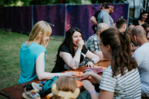Bingo by Quizlabor Zwei Frauen lachen am Tisch, während sie am Elbenwald Festival teilnehmen. Im Hintergrund sind andere Festivalbesucher zu sehen. Die Szene zeigt eine entspannte Atmosphäre mit bunten Accessoires und einem grünen Festivalgelände.