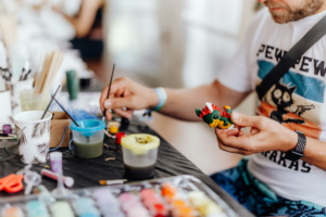 Basteln mit Janina Ein Besucher des Elbenwald Festivals malt ein farbenfrohes Kunstwerk auf einem kleinen Modell, umgeben von Malutensilien und Farben auf einem Tisch. Der kreative Prozess ist Teil der vielfältigen Aktivitäten des Festivals, das Musik und Nerd-Kultur kombiniert.