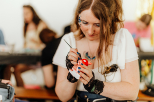 Basteln mit Janina Eine junge Frau mit auffälligem Make-up und schwarzen Spitzenhandschuhen malt sorgfältig eine Figur auf dem Elbenwald Festival. Im Hintergrund sind weitere Teilnehmer mit kreativen Aktivitäten beschäftigt, was die kreative Atmosphäre des Festivals unterstreicht.
