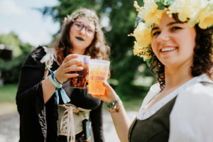 Zwei Frauen in fantasievoller Kleidung mit Blumenkränzen stoßen mit bunten Bechern auf dem Elbenwald Festival an. Die Atmosphäre ist festlich und lebhaft, umgeben von grüner Natur. Ideal für Fans von Musik und nerdiger Kultur.