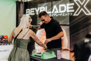 Beyblade Area Zwei Teilnehmer am Beyblade X Stand beim Elbenwald Festival, einer spielt mit einem Beyblade auf einem Spieltisch, während der andere zuschaut. Im Hintergrund ist das Beyblade X Logo deutlich sichtbar.