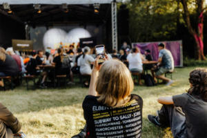 Bingo by Quizlabor Ein Besucher des Elbenwald Festivals sitzt im Gras und filmt die Bühne mit seinem Smartphone. Im Hintergrund sind andere Festivalbesucher zu sehen, die die Aufführung genießen. Die Atmosphäre ist lebhaft und von Vorfreude geprägt.