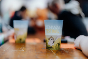 Buntes Trinkglas mit dem Schriftzug "Elbenwald Festival" und einem niedlichen Charakter in einem Bambuswald. Im Hintergrund sind verschwommen Menschen und ein Holztisch zu sehen, die das gesellige Ambiente des Festivals unterstreichen.