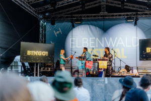 Bingo by Quizlabor Bühne beim Elbenwald Festival mit einem Bingo-Spiel. Eine Moderatorin überreicht einem Teilnehmer eine Karte, während zwei weitere Moderatoren am Tisch stehen. Im Hintergrund ist die Festivalbühne und das Logo des Festivals sichtbar.