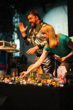 Bingo by Quizlabor Ein Moderator in einem glitzernden Outfit und eine Frau mit blonden Haaren präsentieren eine Auswahl an Sammlerstücken und Merchandise auf der Bühne des Elbenwald Festivals, während das Publikum aufmerksam zuschaut.
