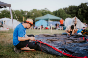 Ein Festivalbesucher beim Aufbauen seines Zeltes auf dem Elbenwald Festival, umgeben von weiteren Zelten und einem grünen Festivalgelände, während die Sonne untergeht. Die entspannte Atmosphäre und die Vorfreude auf das Event sind spürbar.