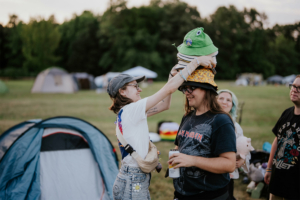 Zwei Festivalbesucher lachen und tragen bunte Hüte auf dem Gelände des Elbenwald Festivals. Im Hintergrund sind Zelte zu sehen, während sie eine entspannte Atmosphäre genießen. Die Szene zeigt die fröhliche und kreative Gemeinschaft des Festivals.