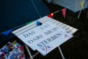 Ein Tisch mit einem auffälligen Schild beim Elbenwald Festival, das den Slogan "Das EWF darf nicht sterben!" zeigt. Im Hintergrund sind Zelt und bunte Wimpel zu sehen, die die festliche Atmosphäre unterstreichen.