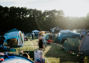 Zeltplatz des Elbenwald Festivals mit bunten Fähnchen, Sonnenstrahlen und Besuchern, die sich zwischen den Zelten bewegen. Die Atmosphäre ist lebhaft und einladend, ideal für Fans von Musik und Nerdkultur.
