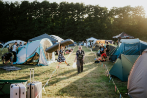 Zeltplatz beim Elbenwald Festival mit Festivalbesuchern, die ihre Ausrüstung aufbauen. Eine Frau trägt eine Luftmatratze, während andere mit Zelten beschäftigt sind. Die Atmosphäre ist lebhaft und freundlich, umgeben von Bäumen und Festivalzelten.