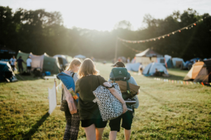 Drei Festivalbesucher gehen mit Rucksäcken und Taschen über das Gelände des Elbenwald Festivals. Im Hintergrund sind Zelte und festliche Lichter zu sehen, während die Sonne am Horizont aufgeht und eine lebhafte Atmosphäre schafft.