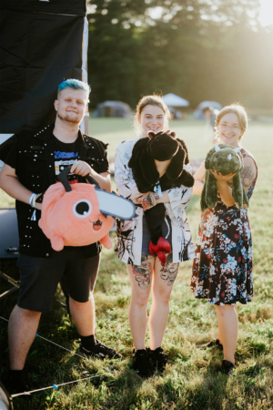 Drei Festivalbesucher halten kuschelige Plüschtiere in der Hand, während sie auf dem Elbenwald Festival stehen. Die Szene zeigt eine fröhliche Atmosphäre mit einem sonnigen Hintergrund und Zelten im Hintergrund, ideal für Fans von Musik und Nerdkultur.