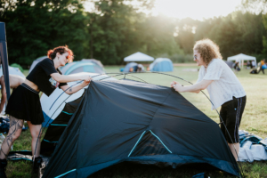 Zwei Festivalbesucherinnen beim Aufbauen eines Zeltes auf dem Elbenwald Festival, umgeben von weiteren Zelten und einer grünen Wiese. Die Sonne scheint, während die Atmosphäre der Veranstaltung eine Mischung aus Musik und Gemeinschaft vermittelt.