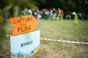Schild mit der Aufschrift "Capture the Flag" auf dem Elbenwald Festival, das auf die Spielfläche "Reitwiese" weist. Im Hintergrund sind Festivalbesucher zu sehen, die an verschiedenen Aktivitäten teilnehmen.