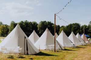 Zeltlager auf dem Elbenwald Festival, mit weißen Tipis in einer grünen Wiese, umgeben von Bäumen und festlicher Beleuchtung. Die Einrichtung umfasst weiße Tische und Stühle, ideal für entspannte Gespräche und gemeinsames Verweilen.
