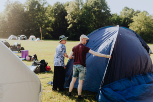 Zwei junge Männer bauen ein Zelt auf dem Elbenwald Festival auf, umgeben von weiteren Zelten und Campingausrüstung auf einem sonnigen Festivalgelände. Die Atmosphäre ist entspannt und freundlich, ideal für Fans von Musik und Nerdkultur.