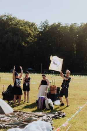 Vier Festivalbesucher feiern auf dem Elbenwald Festival, während einer eine Flagge schwenkt. Die Szene zeigt eine fröhliche Atmosphäre mit Zelten und Gepäck im Hintergrund, umgeben von bunten Wimpeln und grünem Gras.