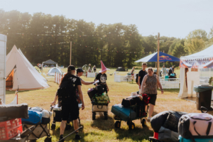 Festivalbesucher beim Elbenwald Festival transportieren ihre Ausrüstung in Bollerwagen über das Gelände. Im Hintergrund sind Zelte und Stände zu sehen, während die Sonne scheint und eine fröhliche Atmosphäre herrscht.