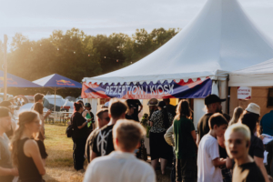 Menschenmenge vor dem Empfangs- und Kiosk-Zelt beim Elbenwald Festival, umgeben von Zelten und Red Bull-Schirmen, in einer sonnigen Abendstimmung. Besucher warten in einer lebhaften Atmosphäre auf Informationen und Snacks.