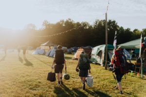 Zwei Festivalbesucher mit Rucksäcken und Gepäck gehen auf einem sonnigen Gelände zum Elbenwald Festival, umgeben von Zelten und bunten Lichtern, während die morgendliche Sonne eine gemütliche Atmosphäre schafft.