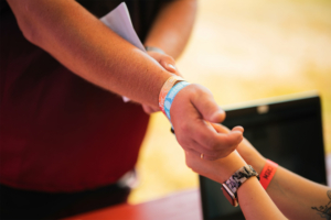 Hände, die ein Festivalarmband anlegen, während im Hintergrund ein Laptop sichtbar ist. Die Szene zeigt die Anmeldung oder den Zugang zum Elbenwald Festival, das Musik und Nerdkultur kombiniert.
