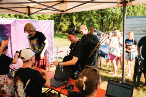 Besucher des Elbenwald Festivals stehen an einem Informationsstand, während ein Teilnehmer mit einem großen Rucksack und Ausrüstung freundlich empfangen wird. Im Hintergrund sind weitere Festivalbesucher und ein bunter Veranstaltungsbanner zu sehen.