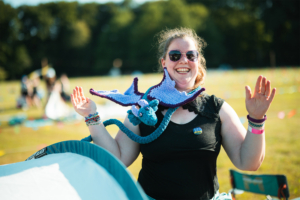 Lächelnde Frau auf dem Elbenwald Festival mit einem gehäkelten Drachen um den Hals, umgeben von Festivalatmosphäre und bunten Dekorationen. Sie trägt eine schwarze Bluse und zahlreiche Armbänder, während sie fröhlich in die Kamera winkt.