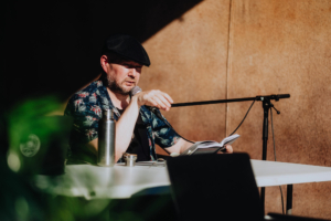 Christian von Aster Ein Mann mit einer Schirmmütze und einem bunten Hemd sitzt an einem Tisch und hält ein Mikrofon, während er aus einem Buch liest. Im Hintergrund sind Pflanzen und ein Laptop zu sehen, die die entspannte Atmosphäre des Elbenwald Festivals unterstreichen.