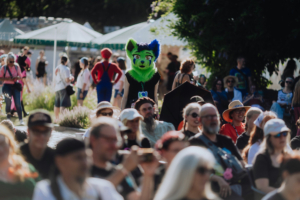 Bunt gekleidete Festivalbesucher auf dem Elbenwald Festival, darunter eine Person im auffälligen Fursuit mit grünen und blauen Akzenten, während andere in verschiedenen Kostümen und alltäglicher Kleidung im Hintergrund stehen.