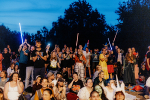 Menschenmenge beim Elbenwald Festival, die enthusiastisch mit Lichtschwertern und Handys feiert. Verschiedene Kostüme und fröhliche Gesichter unter einem Abendhimmel, der die festliche Stimmung des einzigartigen Events widerspiegelt.
