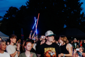 Zwei Festivalbesucher heben leuchtende Lichtschwerter in die Luft, während die Menge beim Elbenwald Festival feiert. Die Atmosphäre ist lebhaft und voller Energie, umgeben von begeisterten Fans und bunten Lichtern.