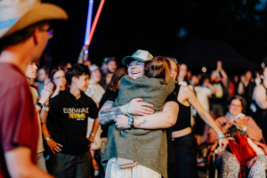 Zwei Frauen umarmen sich herzlich auf dem Elbenwald Festival, umgeben von fröhlichen Festivalbesuchern. Im Hintergrund sind bunte Lichter und die lebendige Atmosphäre eines einzigartigen Musik- und Nerd-Events zu sehen.