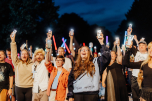 Eine begeisterte Menschenmenge hebt ihre Smartphones mit leuchtenden Displays und Lichtschwertern in die Luft während des Elbenwald Festivals, das eine lebendige Mischung aus Musik und Nerd-Kultur bietet. Die Atmosphäre ist voller Freude und Gemeinschaft.
