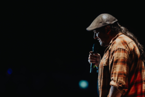 Ein männlicher Redner mit einem Hut und einer karierten Jacke hält ein Mikrofon auf der Bühne beim Elbenwald Festival. Die Atmosphäre ist energiegeladen, mit einem dunklen Hintergrund und einem schwachen Lichtschein im Bild.