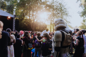 Ein Stormtrooper steht inmitten einer begeisterten Menge beim Elbenwald Festival, umgeben von bunten Kostümen und einer lebhaften Atmosphäre. Im Hintergrund ist die Bühne zu sehen, während die Sonne durch die Bäume strahlt.