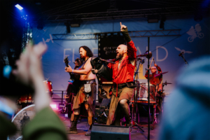 Corvus Corax Zwei Musiker in historischer Kleidung treten auf der Bühne des Elbenwald Festivals auf. Der Gitarrist spielt leidenschaftlich, während der Sänger das Publikum begeistert. Im Hintergrund sind Schlaginstrumente und weitere Bandmitglieder sichtbar.