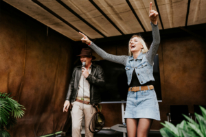 Cosplay Catwalk Zwei begeisterte Teilnehmer auf der Bühne des Elbenwald Festivals, einer Mischung aus Musikfestival und Convention. Die Frau in einem Jeansoutfit jubelt, während der Mann in Lederjacke und Hut spricht. Die Atmosphäre ist lebhaft und einladend.